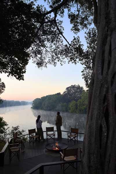 Guests on the riverbank beneath an ebony tree at dusk